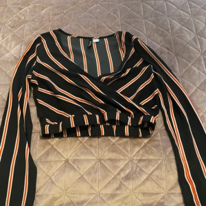 Randig omlottopp från H&M Divided, S - Säljer en croppad omlottopp från H&M Divided med svarta, orangea och vita ränder. Toppen har långa ärmar, v-ringning och resår i midjan. Perfekt för dig som gillar trendiga och färgstarka plagg med snygg passform.