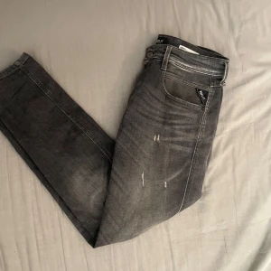 Replay Anbass grå jeans med slitningar - Säljer ett par Replay Anbass jeans i grå tvätt med snygga slitningar framtill. Jeansen har klassisk femficksdesign, smal passform och är tillverkade i stretchig bomullsdenim för extra komfort. Perfekta för dig som gillar en avslappnad men trendig look. Använda 4-5 gånger, köpta för 2000, byten funkar också