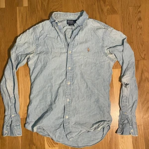 Polo Ralph Lauren skjorta - Säljer en ljusblå skjorta från Polo Ralph Lauren i slim fit. Skjortan har klassisk krage, långa ärmar med manschett och knappar framtill. Materialet är mjuk bomull och den har den ikoniska broderade loggan på bröstet. Perfekt för dig som gillar stilren och enkel look. Typ tunnt jeanstyg.