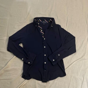 Mörkblå skjorta från Polo Ralph Lauren - Klassisk mörkblå skjorta från Polo Ralph Lauren med långa ärmar och vita knappar. Insidan av kragen har snygga ränder i rött, vitt och blått samt små broderade lejon i gult. Skjortan är i mjuk bomull och har broderad logga på bröstet.