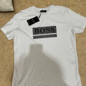 Vit t-shirt från Hugo Boss - Snygg vit t-shirt från Hugo Boss med stort svart logotryck på bröstet. Perfekt för dig som gillar stilrena och trendiga plagg med tydlig branding.