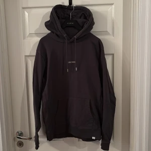 Svart hoodie från Les Deux - Snygg svart hoodie från Les Deux med minimalistisk logga på bröstet. Tröjan har huva med snörning och en stor magficka framtill. Perfekt för en chill och stilren look.