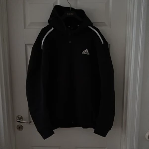 Svart hoodie från Adidas med huva - Svart hoodie med dragkedja från Adidas med vit logga på bröstet och vita ränder längs axlarna. Tröjan har huva, lång ärm och klassisk sportig look. Perfekt för dig som gillar streetwear eller sportig stil.