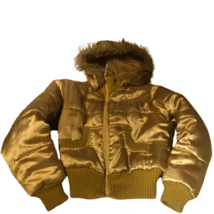 Guldig y2k pufferjacka med huva från Original Denim - Storlek XL men jag skulle säga att den sitter som en s/xs som nån som alltid har xxs. Den är cropped så. Säljer en guldig pufferjacka från Original Denim med huva och fuskpäls. Jackan har dragkedja, ribbade muddar och nederkant samt två fickor framtill. Perfekt för kyligare dagar och ger en cool streetstil. Väldigt fin till mcbling/y2k stil 