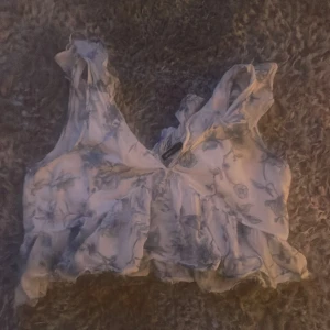 Blommig volang topp med knyt - Supersöt blommig topp med volangdetaljer och knytning fram. Ljusblått och vitt mönster, ärmlös och croppad modell. 