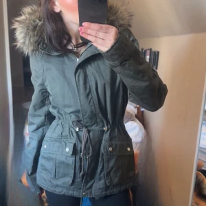 Grön parkajacka med fuskpäls - Snygg grön parkajacka med dragsko i midjan och stor huva med fuskpäls. Jackan har dragkedja och knappar framtill samt stora fickor. Perfekt för kyligare dagar.