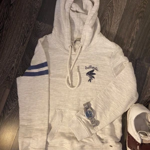 Ljusgrå hoodie med broderi - Säljer en ljusgrå knittad hoodie med broderad text 'Authentic' och indianmotiv på bröstet. Tröjan har huva med dragsko, känguruficka och två blå ränder på ena ärmen. Perfekt för en avslappnad stil. Ny pris 3350kr