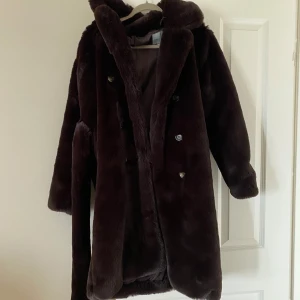 Mörkbrun pälsjacka från Urbancode - Säljer en lång, mörkbrun pälsjacka från Urbancode med bälte i midjan. Jackan har dubbelknäppning med knappar och är helfodrad. Perfekt för kalla dagar när du vill vara både varm och snygg! (Jackan är fuskpäls!)