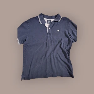 Mörkblå pikétröja från Champion - Säljer en mörkblå pikétröja från Champion med vit logga på bröstet och vita detaljer på kragen. Tröjan har korta ärmar och knäppning framtill. Perfekt för en sportig och avslappnad stil.