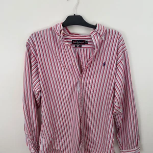 Röd- och vitrandig skjorta från Ralph Lauren - Säljer en klassisk skjorta från Ralph Lauren med röda och vita ränder samt blå logga broderad på bröstet. Skjortan har lång ärm och knäppning framtill. Perfekt för dig som gillar tidlös stil och vill ha något från ett ikoniskt märke.