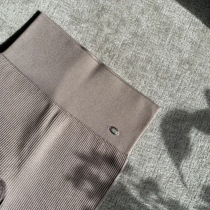 Ribbade beige cykelshorts från DOM - Säljer ett par ribbade beige cykelshorts med hög midja och stretchigt mjukt material. Perfekta för träning eller en avslappnad stil. De har en diskret logga framtill i midjan och är knälånga. 