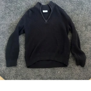 Svart stickad tröja med krage från Grunt - Säljer en svart stickad tröja från Grunt i storlek S. Tröjan har lång ärm, ribbad struktur och en liten krage med v-ringad öppning. Perfekt för lager-på-lager och enkel att matcha med olika outfits.