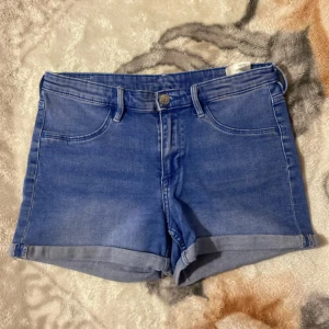 Blå jeansshorts från H&M - Snygga blå jeansshorts från H&M med skinny fit och klassisk femficksdesign. Shortsen har uppvikta benslut, bälteshällor och knappgylf. Perfekta för sommardagar och enkla att matcha med olika toppar.