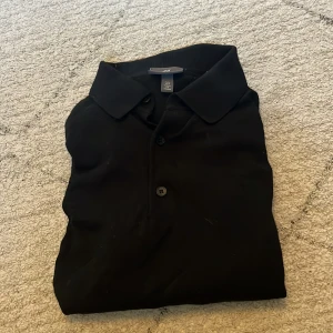 Svart pikétröja från H&M - Säljer en svart långärmad pikétröja från H&M i slim fit-modell. Tröjan har klassisk krage och knäppning framtill med två knappar. Perfekt för en stilren och enkel look.