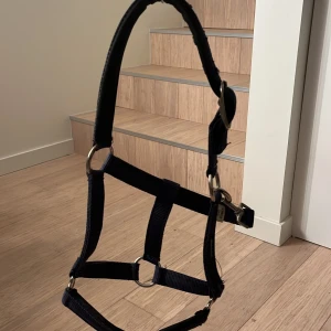 Mörkblå/Marinblå grimma - Säljer en marinblå grimma som är storleken ponny men har ett justerbart spänner på sidan. Grimman har använts och är lite slitet på insidan av grimman där spännet går igenom ( skickar bild om du vill ) och så är även den lite rostig på båda metallerna längst fram vid mulen och även på spännet. Annars en väldigt fin grimma som man kan få tvättade innan den skickas. Grimman är ifrån märket Hasbo sport som man såg på bild 2. Pris kan diskuteras 