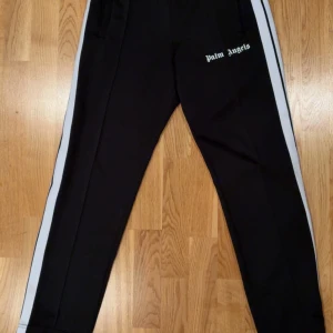 Svarta trackpants från Palm Angels med vita ränder - Svarta trackpants från Palm Angels med vita sidoränder och logga framtill. Byxorna har normal passform och är gjorda i polyester. Perfekta för en streetwear-look. Skick 10/10, kan även skicka fler bilder om man vill det. Nypris 5870kr. 2500kronor är mitt pris utifrån det perfekta skicket och utifrån marknadspriset är det mer än 50% billigare! Tveka inte på fråga vad som helst