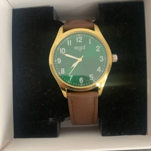 Grön klocka med brunt skinnarmband från Regal - Stilren klocka från Regal med grön urtavla, guldfärgat boett och brunt skinnarmband. Klockan har tydliga vita siffror och klassisk design som passar till många tillfällen.