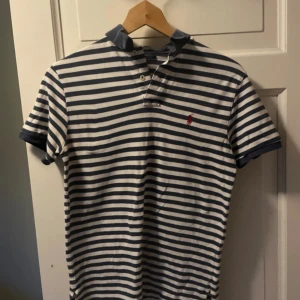 Randig pikétröja från Polo Ralph Lauren - Säljer en klassisk blåvit randig pikétröja från Polo Ralph Lauren. Tröjan har korta ärmar, krage och knappar framtill samt den ikoniska loggan broderad på bröstet. Tillverkad i 100% bomull för en skön känsla.