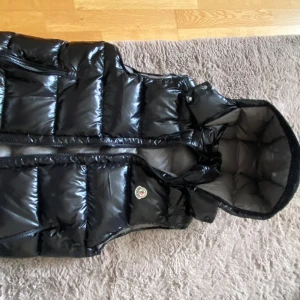 Svart dunväst från Moncler med huva - Säljer en svart, glansig dunväst från Moncler med avtagbar huva och dragkedja framtill. Västen har klassisk Moncler-logga på bröstet och ett unikt serietidningsfoder på insidan.