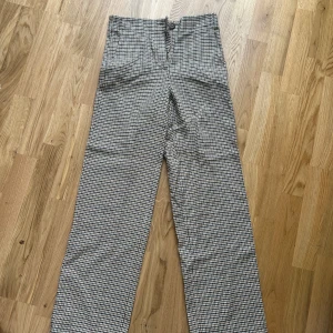 Rutig kostymbyxa från Massimo Dutti - Snygga kostymbyxor från Massimo Dutti i ett klassiskt rutigt mönster i svart, vitt och brunt. Byxorna har normal passform, raka i benen och mellan hög midja och stängs med knapp och dragkedja. Perfekta för en stilren look.