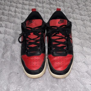 Nike sneakers i svart och rött - Säljer ett par klassiska Nike sneakers i svart och rött med vit sula. Skorna har snörning, rund tå och den ikoniska Nike-loggan på sidan. Perfekta för dig som gillar streetstyle och vill ha en snygg färgkombo.   Storlek 38 