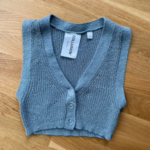 Grå stickad väst från COLLUSION - Grå stickad väst från COLLUSION med v-ringning och tre knappar framtill. Modellen är croppad och har en mjuk, ribbad struktur som ger en avslappnad och trendig look.