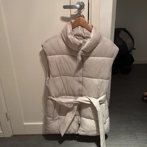 Ljusbeige dunväst med bälte från H&M XS - Säljer en ljusbeige dunväst från H&M med hög krage och bälte i midjan. Västen är quiltad och har en stilren look, perfekt för lager-på-lager. Passar dig som gillar en enkel men trendig stil. Den är aldrig andvänd och ny skick. Den är i strl XS (160)