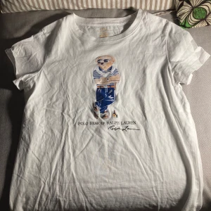Vit t-shirt med Polo Bear från Ralph Lauren - Vit t-shirt från Ralph Lauren med ikoniska Polo Bear-trycket framtill. Björnen har blå byxor och randig tröja, vilket ger en cool och klassisk look. Rund halsringning och korta ärmar. Perfekt för dig som gillar stilrena och lekfulla plagg.