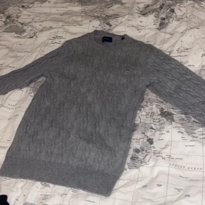 Grå kabelstickad tröja från Gant - Säljer en klassisk grå kabelstickad tröja från Gant med rund halsringning och broderad logga på bröstet. Tröjan har långa ärmar och breda ribbade muddar vid ärmslut och nederkant. Perfekt för lager-på-lager-stil.