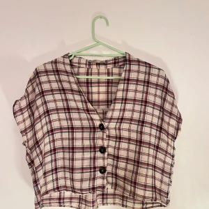 Rutig kortärmad blus med knappar - Säljer en rutig blus i beige, rött och svart med v-ringning och knappar framtill. Blusen är kortärmad och har en avslappnad passform!