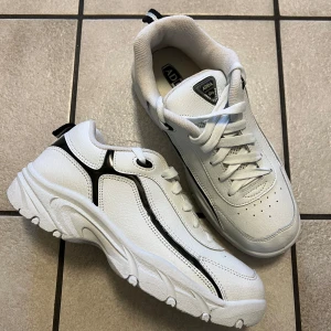 Vita sneakers från Adda med svarta detaljer - Snygga vita sneakers från Adda med svarta detaljer och sportig design. Skorna har snörning, rund tå och en rejäl, mönstrad sula för bra grepp. Perfekta för en stilren och sportig look.