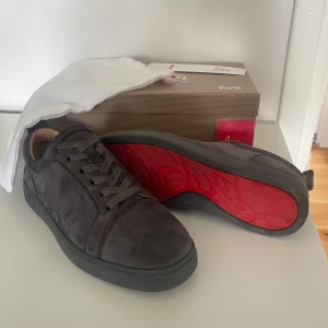 Mörkgrå skor från Christian Louboutin - Säljer ett par mörkgrå sneakers i mocka från Christian Louboutin med klassisk röd sula. Skorna har rund tå, platt sula och snörning. Insidan är ljusbrun och de kommer med originalkartong och dustbag. Säljer för att de aldrig användas, pris går att diskutera vid snabb affär🤩