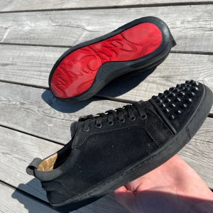 Svarta Louboutins med nitar och röd sula - Säljer ett par svarta sneakers från Christian Louboutin med mattsvarta nitar på tån och ikonisk röd sula. Skorna har snörning och är tillverkade i mocka med svarta detaljer. Perfekta för dig som vill sticka ut med din stil. En nit på vänstra skon har gått av (se sista bilden)