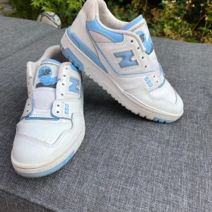 New Balance 550 sneakers i vitt och ljusblått - Säljer ett par New Balance 550 sneakers i vitt med ljusblå detaljer. Skorna har klassisk siluett, snörning och platt sula. Perfekta för dig som gillar sportig och stilren look. Märket syns tydligt på sidan och bak.