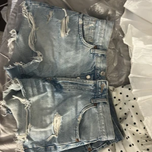 Ljusblå jeansshorts med fransar - Säljer ett par ljusblå jeansshorts med slitna detaljer och fransiga benslut. Shortsen har klassisk femficksdesign, knappgylf och hög midja. Perfekta för en avslappnad stil. Från Zara! Skriv vid intresse!! 