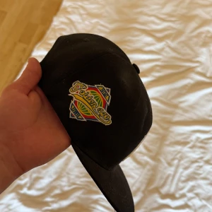 Svart keps med färgglad broderad logga - Svart keps med en färgglad broderad logga framtill där det står 'Pormayor'. Kepsen har böjd skärm och justerbar snapback baktill. Perfekt accessoar för en cool och avslappnad stil.