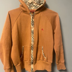 Beige hoodie från Burberry med rutiga detaljer - Säljer en beige hoodie från Burberry med klassiskt rutigt foder i huvan och längs dragkedjan. Jackan har fickor framtill och en liten röd Burberry-logga på bröstet. Perfekt för dig som gillar streetwear med lyxig känsla.