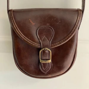 Brun axelväska från Ann Nichols äkta läder - Säljer en klassisk brun axelväska i skinn från Ann Nichols. Väskan har ett spänne framtill med metall-detalj och en justerbar axelrem. Baksidan har ett litet fack med dragkedja. Perfekt för dig som gillar vintage och tidlös stil.