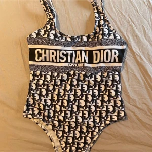 Svartvit baddräkt från Christian Dior - Säljer en snygg svartvit baddräkt från Christian Dior med tydlig logotyp framtill och klassiskt Dior-mönster över hela plagget. Baddräkten har breda axelband och rundad halsringning. Perfekt för sommaren!