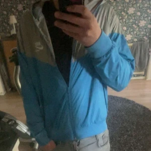 Blå och grå vindjacka från Nike - Säljer en blå och grå vindjacka från Nike med huva och dragkedja framtill. Jackan har lång ärm och sportig look, perfekt för utomhusaktiviteter. Nike-logga på bröstet och ribbade muddar vid ärmslut och nederkant.