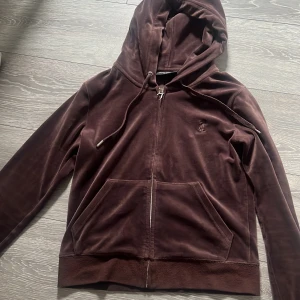 Brun velour hoodie från Juicy Couture - Mysig brun hoodie i velour från Juicy Couture med dragkedja framtill och huva. Tröjan har fickor på magen och snörning i huvan. Perfekt för en avslappnad och trendig look.