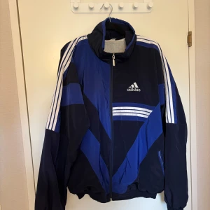Blå och svart vindjacka från Adidas - Retro vindjacka från Adidas i blått och svart med vita detaljer och klassiska ränder på ärmarna. Jackan har dragkedja framtill, hög krage och två fickor. Perfekt för en sportig stil.