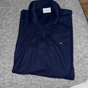 Mörkblå pikétröja från Tommy Hilfiger - Säljer en klassisk mörkblå pikétröja från Tommy Hilfiger med liten logga på bröstet. Tröjan har krage och knappar framtill, perfekt för en stilren look.