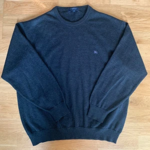 Burberry Tröja Merinoull - Säljer en stilren mörkgrå tröja från Burberry i 60% merinoull, självklart äkta. Tröjan har rund halsringning och diskret broderad logga på bröstet. Perfekt för dig som gillar en klassisk och enkel design. Storlek Medium (7).