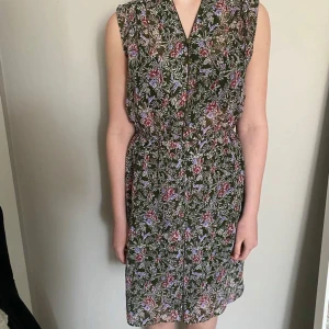 Grön blommig klänning med knappar - Säljer en grön blommig klänning med röda och lila blommor. Klänningen är ärmlös, har knappar framtill och resår i midjan för en bekväm passform. Perfekt för varma dagar och har en luftig känsla.