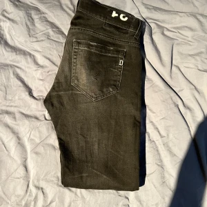 Svarta jeansbyxor från Dondup - Säljer ett par svarta jeansbyxor från Dondup med klassisk femficksdesign och raka ben. Jeansen har diskreta slitningar och en liten Dondup-logga på bakfickan. Perfekta för dig som gillar en stilren look.