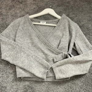 Grå omlottstickad tröja från Kappahl - Mysig grå stickad omlott-tröja från Kappahl med knyt i sidan. Tröjan har lång ärm och är croppad i modellen, perfekt för lager-på-lager. Enkel och stilren design som passar till mycket. Aldrig använd.