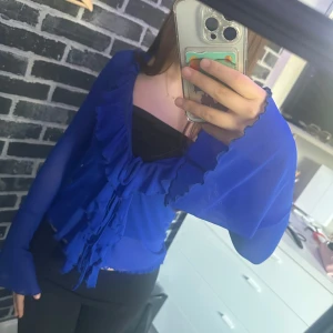 Kornblå volangblus med knyt - Säljer en kornblå blus med långa ärmar och volangdetaljer längs v-ringningen. Blusen har knyt framtill och är tillverkad i ett lätt, transparent material. Perfekt för att lyfta en outfit med lite extra färg.