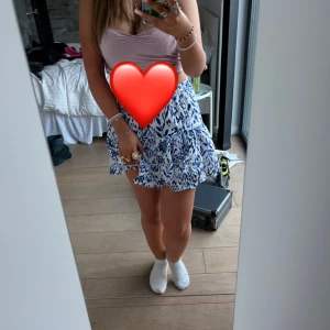 Blåvit volangkjol med mönster - Säljer en kort blåvit kjol med volanger och ett snyggt abstrakt mönster. Kjolen har en luftig känsla och passar perfekt för varmare dagar. Resår i midjan för extra komfort.