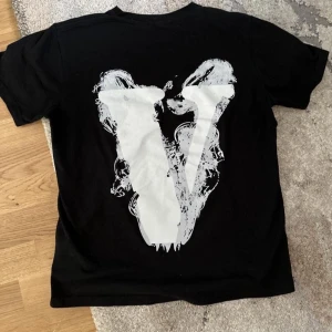 Svart v-lone T-shirt  - Cool svart t-shirt från Vlone med stor vit V-logga på ryggen och unik ängel- och textgrafik på bröstet. T-shirten har rund hals och korta ärmar, perfekt för en streetwear-look. Nästan helt oanvänd 